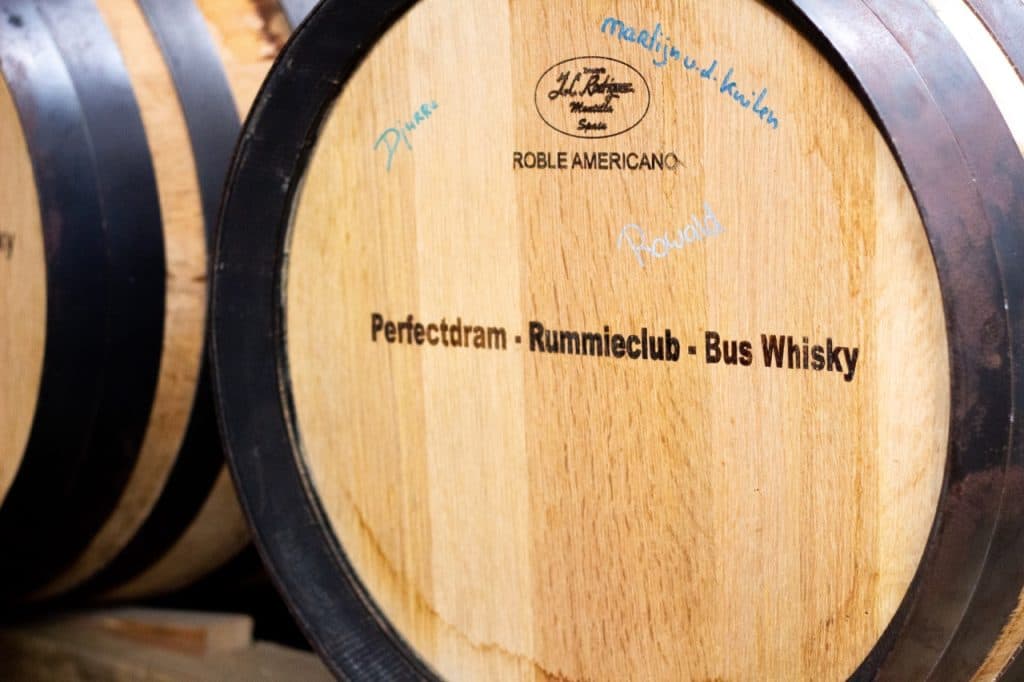 Een close-up van een houten vat met het etiket "Perfectdram - Rummieclub - Bus Whisky" met "ROBLE AMERICANO" en andere handgeschreven notities, gespot bij Uitboeren in Den Bosch. De houtnerf en zwarte metalen banden zijn duidelijk zichtbaar.