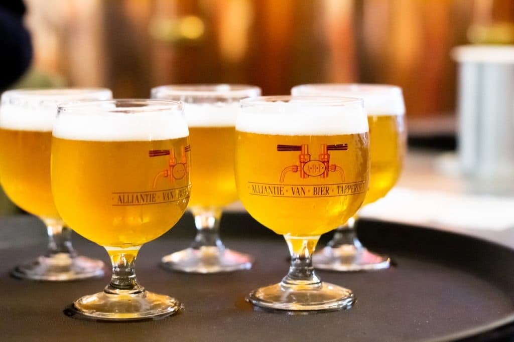 Vijf met schuimend goudkleurig bier gevulde glazen met steel rusten op een rond zwart dienblad. De glazen, gemerkt met Uitboeren Den Bosch in rode tekst en logo, steken af tegen de zachte achtergrond van warme houttinten en heldere verlichting.