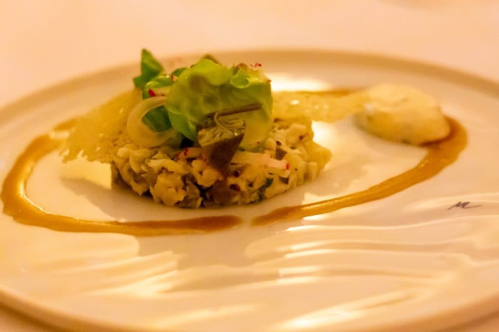Een gastronomisch gerecht op een wit bord, geïnspireerd door Uitboeren in Den Bosch, met een klein hoopje fijngesneden groenten met daarop sla en knapperig garnituur. Een bruine saus wordt kunstig omgeroerd, naast een toef romig wit schuim of puree.