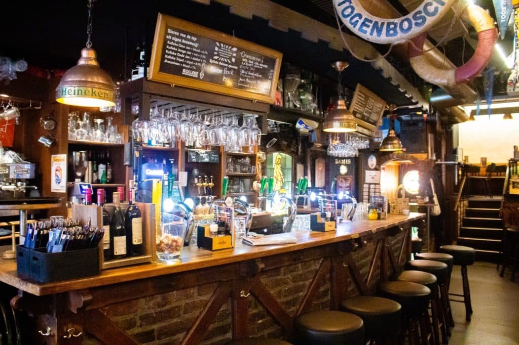 Een gezellige bar in Den Bosch met een houten toonbank met krukken, verschillende flesjes en biertaps. Boven hangen glazen en koperen lampen, terwijl de krijtbordmenu's en Uitboeren decoraties een warme, uitnodigende sfeer creëren.