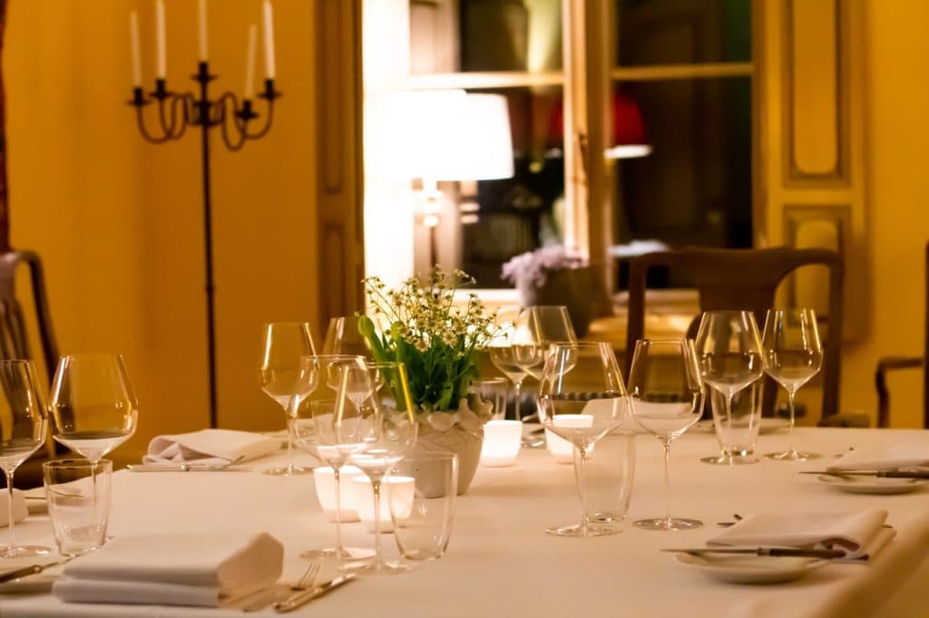 Een warm verlichte eetkamer in Den Bosch heeft een wit tafelkleed, netjes gerangschikte wijnglazen, borden en servetten. Een kleine potplant is het middelpunt, met stoelen, een kandelaar en een gloeiende lamp bij grote ramen die de uitboeren-sfeer compleet maken.