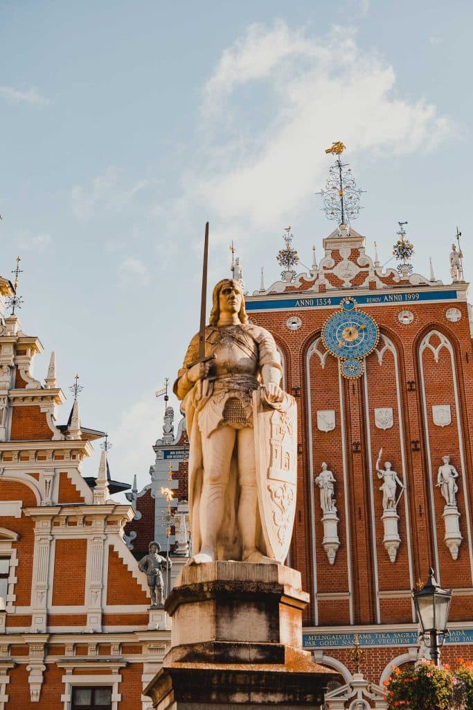 Een stenen standbeeld van een middeleeuwse ridder met een zwaard en schild staat op een sokkel in Riga, Letland. Daarachter staat een sierlijk gebouw van rode baksteen met witte versieringen, een blauwe klok, beelden en gouden accenten onder een heldere halfbewolkte hemel.