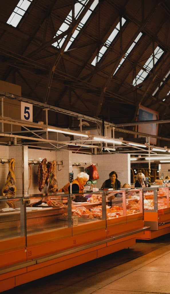Een helder verlichte marktkraam in Riga toont verschillende stukken vlees achter glas. Twee verkopers helpen klanten, met groot hangend vlees op de achtergrond. De Letland markt heeft een hoog, industrieel plafond en is gemarkeerd met een blauw "5" bord.