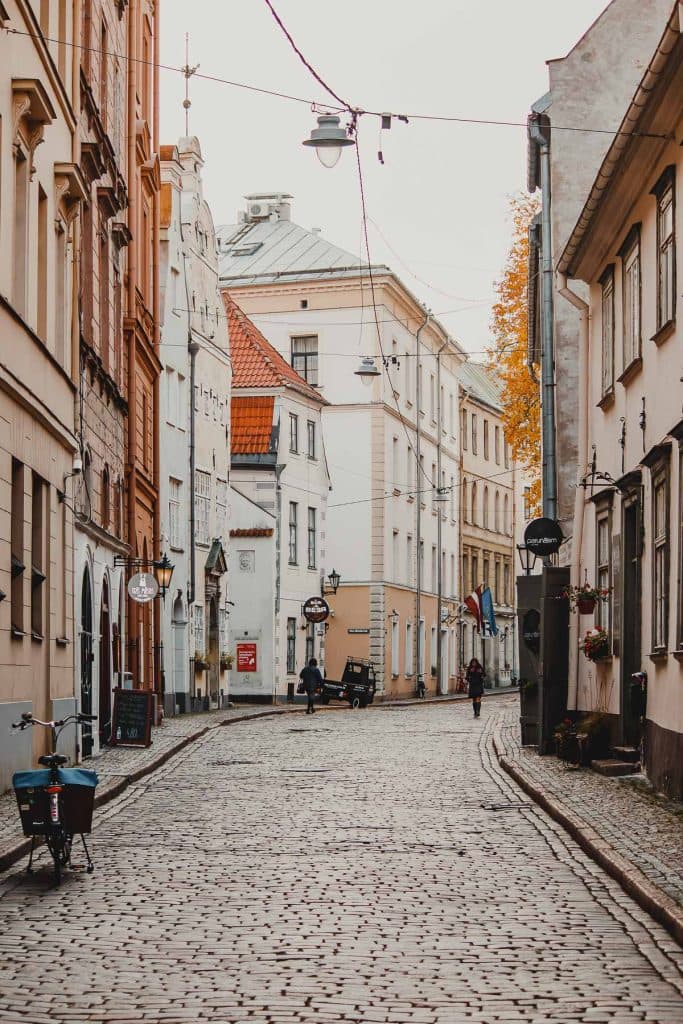 Een geplaveide straat in Riga, Letland, omzoomd met historische gebouwen, fietsen en uithangborden. Een paar mensen lopen in de verte, genietend van het rustige stadsgezicht en de herfstbladeren aan een nabijgelegen boom.