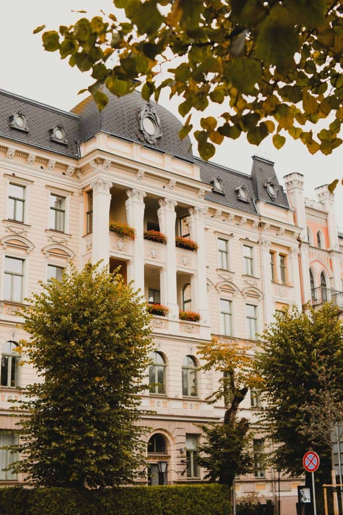 Elegant historisch gebouw met sierlijke zuilen en balkons versierd met bloembakken, gedeeltelijk aan het oog onttrokken door lommerrijke bomen in Riga; rechtsonder is een bord met een parkeerverbod te zien - een charmant gezicht tijdens een stedentrip naar Letland.