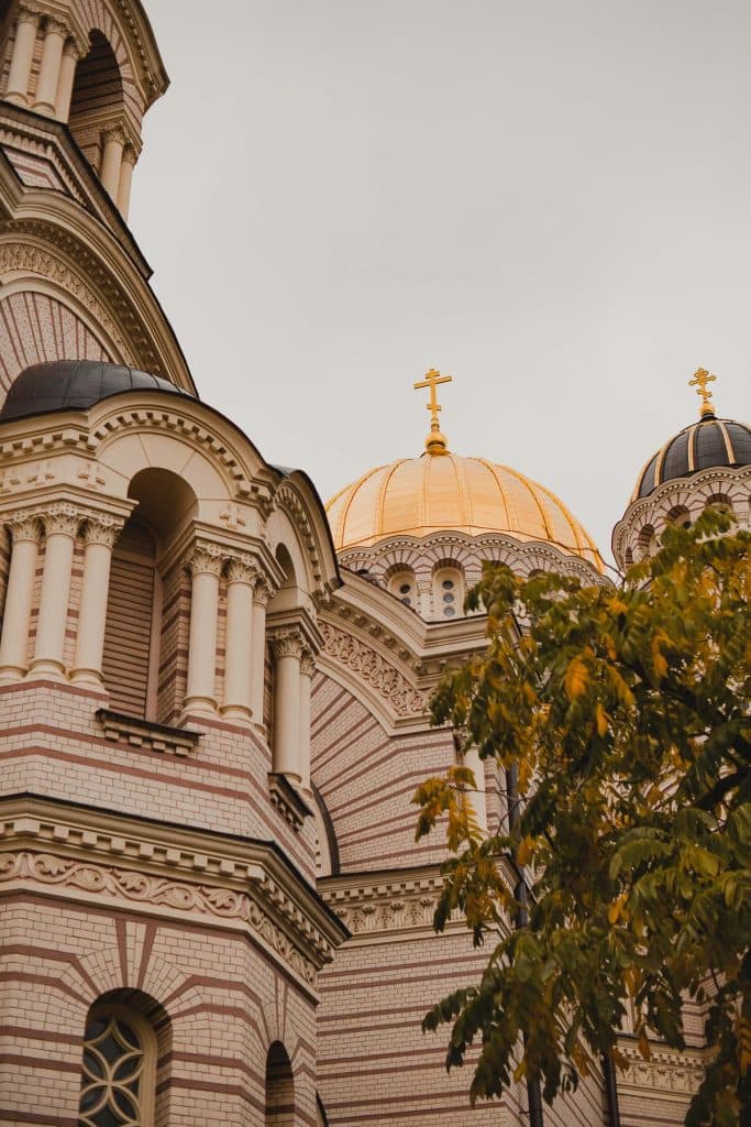 Close-up van een sierlijke kerk in Riga met roze en wit gestreept metselwerk, boogramen en een gouden koepel met een kruis erop, gedeeltelijk verborgen door groene takken - perfect voor een Letland citytrip.