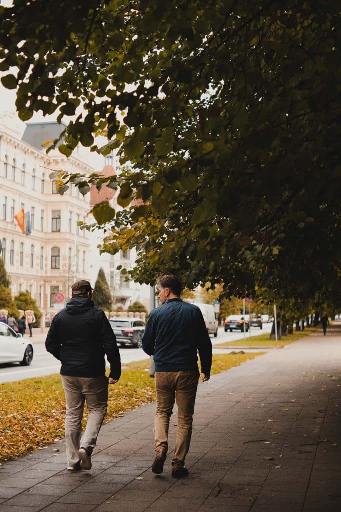 Twee mensen lopen over een met bomen omzoomd trottoir in Riga op een herfstdag, met afgevallen bladeren onder hun voeten. Auto's en historische gebouwen versterken de charme van deze Letlandse citytrip, zichtbaar net voorbij de straat links van hen.
