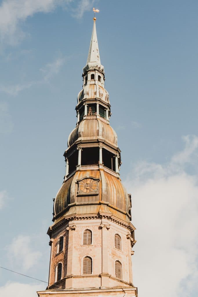 Een hoge, sierlijke kerktoren met een koperen koepel en klok steekt uit boven de skyline van Riga, perfect voor een stedentrip Letland. Het bouwwerk heeft meerdere verdiepingen, decoratieve details en steekt af tegen een blauwe lucht met verspreide wolken.