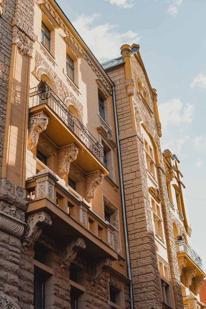 Zonnig uitzicht op een sierlijk, historisch gebouw in Riga, Letland, met decoratieve balkons, gebeeldhouwde stenen details en grote ramen, gefotografeerd vanuit een lage hoek tegen een gedeeltelijk bewolkte blauwe lucht - de perfecte aanblik tijdens een stedentrip.