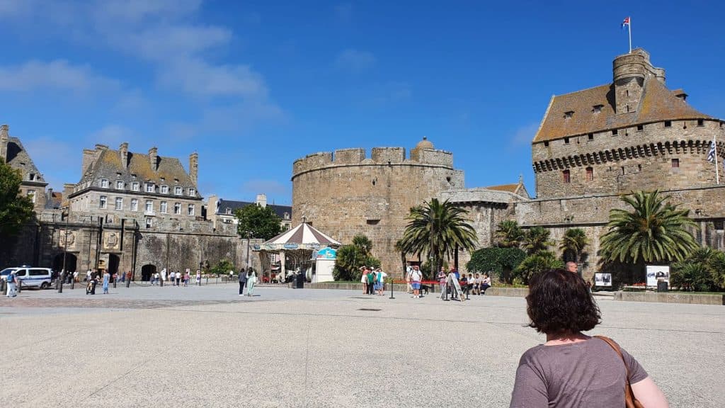 Een persoon met kort bruin haar staat op een groot open plein, tegenover een oud stenen fort met torens en palmbomen, dat de sfeer oproept van een roadtrip door midden frankrijk op een zonnige dag onder een strakblauwe hemel.