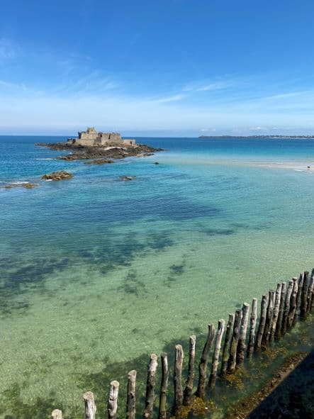 Een stenen fort ligt op een rotsachtig eilandje in een turkooisblauwe zee, gezien vanaf de kust - een iconisch gezicht voor elke roadtrip door midden Frankrijk. Houten palen omzomen het zandstrand en het heldere water onthult ondergedompelde rotsen onder een helderblauwe hemel.