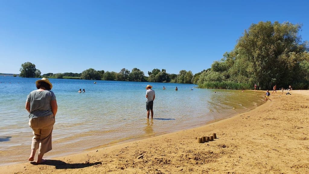 Mensen genieten van een zonnige dag op een zandstrand aan een meer in Frankrijk; een persoon in een gestreept shirt en hoed loopt over de oever, terwijl anderen waden of zwemmen, omringd door groene bomen onder een heldere hemel - een perfecte stop tijdens een roadtrip door midden frankrijk.