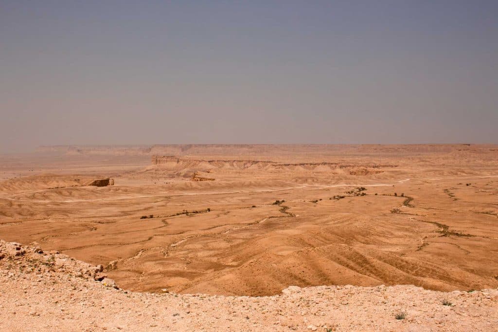 Een uitgestrekt, dor woestijnlandschap strekt zich in de verte uit onder een heldere, wazige hemel in de buurt van Riyad, met rotsplateaus en droge, gebarsten aarde die de ruige schoonheid van Saoedi-Arabië oproepen.