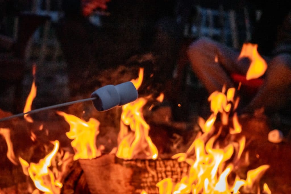 Een marshmallow op een stokje wordt boven een kampvuur met fel oranje vlammen gehouden. Op de achtergrond zitten mensen rond het vuur bij een Ardense tiny house, hun benen zichtbaar, waardoor een gezellige buitensfeer ontstaat in de schemering.