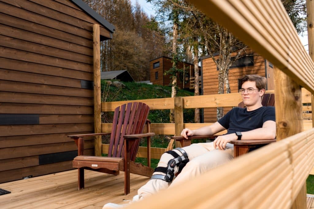 Een jongere met een bril en een kniebrace zit op een houten terras in een buitenstoel, omringd door kleine huisjes en groen, en geniet van een vredig glamping-moment.