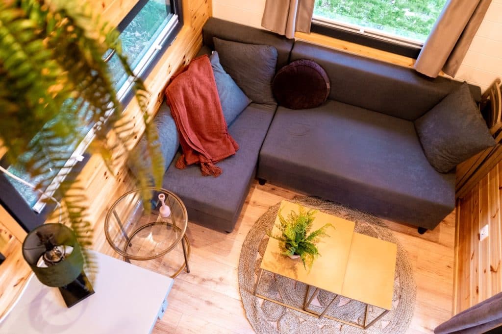 Een gezellige woonkamer in een klein huisje met een L-vormige grijze bank, een rode deken, ronde en vierkante kussens, een kleine gouden salontafel met een plant in potten en grote ramen die natuurlijk licht binnenlaten. Perfect voor glamping binnenshuis.
