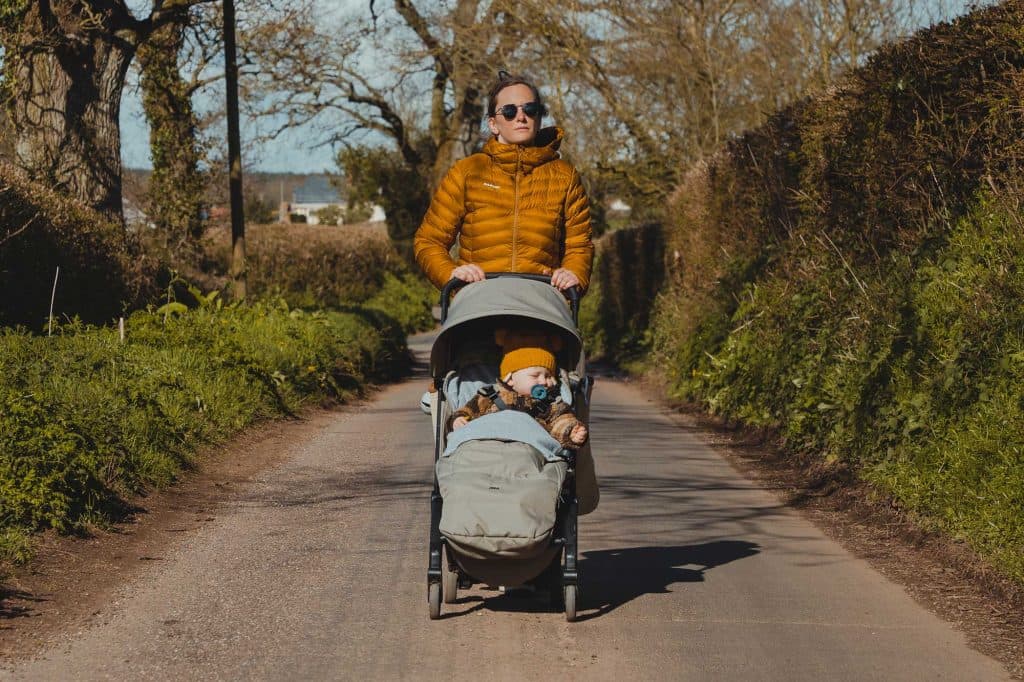 Een persoon met een zonnebril en een geel hesje duwt een kinderwagen met twee jonge kinderen over een landelijk pad met bomen en struiken op een zonnige dag.