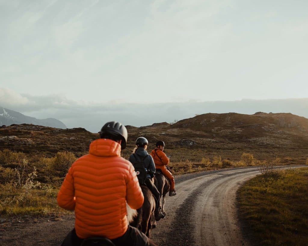 Drie mensen met helmen en oranje jassen rijden op paarden over een gebogen zandweg door een schilderachtig, heuvelachtig Scandinavisch landschap onder een bewolkte hemel, die de wilde schoonheid van Lofoten in Noorwegen oproept.
