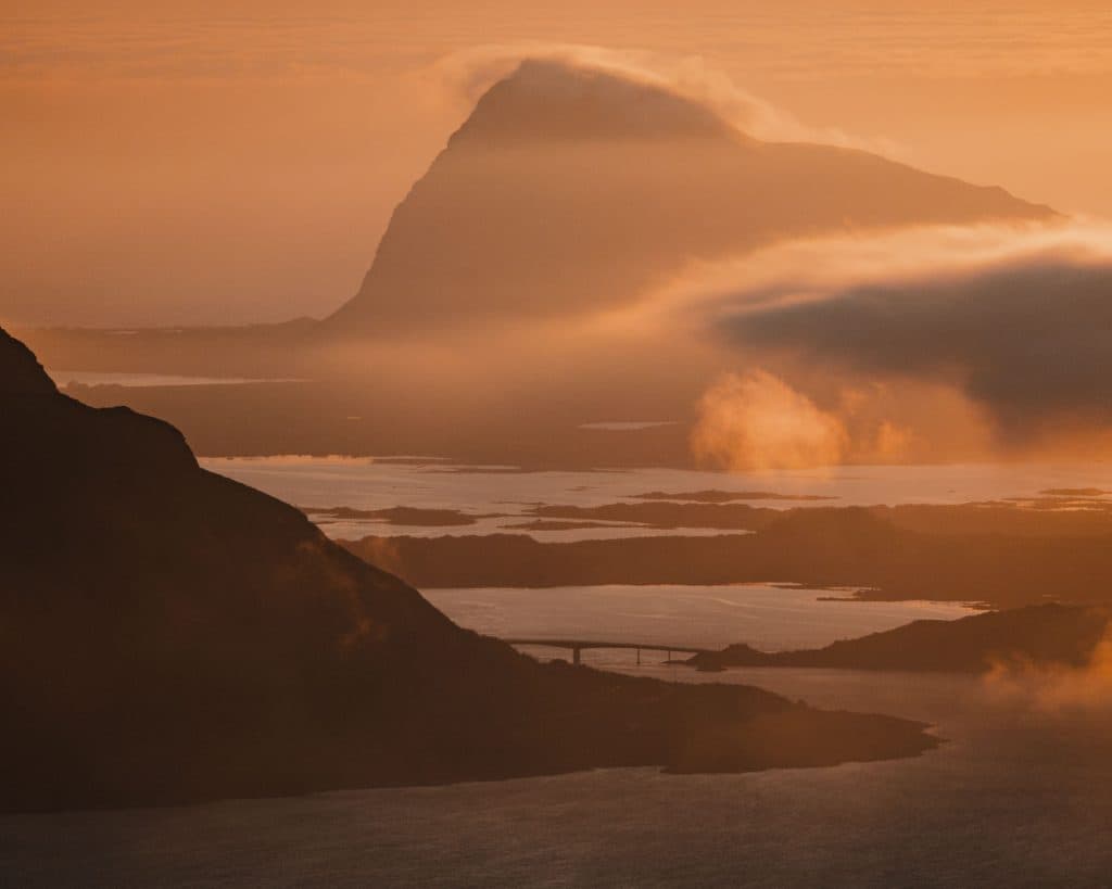 Een mistige Noordse berg rijst op boven kalm water tijdens zonsondergang, met zacht oranje licht en lage wolken die het landschap gedeeltelijk verduisteren. In de verte is vaag een brug te zien die de serene schoonheid van Lofoten oproept.