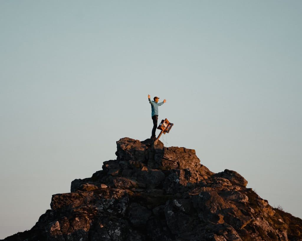 Een persoon staat op de top van een rotsachtige heuvel op de Lofoten, de armen triomfantelijk geheven naast een opgevouwen stoel en rugzak, onder een heldere noorderlucht bij zonsondergang of zonsopgang.