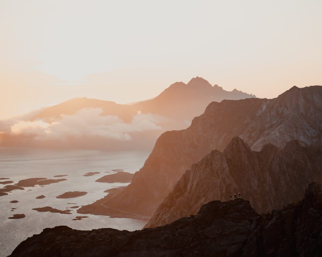 Rotsachtige bergen en eilanden bij zonsondergang met zacht goudkleurig licht, mistige wolken en kalm water eronder; silhouetten van een paar mensen staan op een klif op de voorgrond, een beeld van de serene nordic schoonheid van Lofoten.