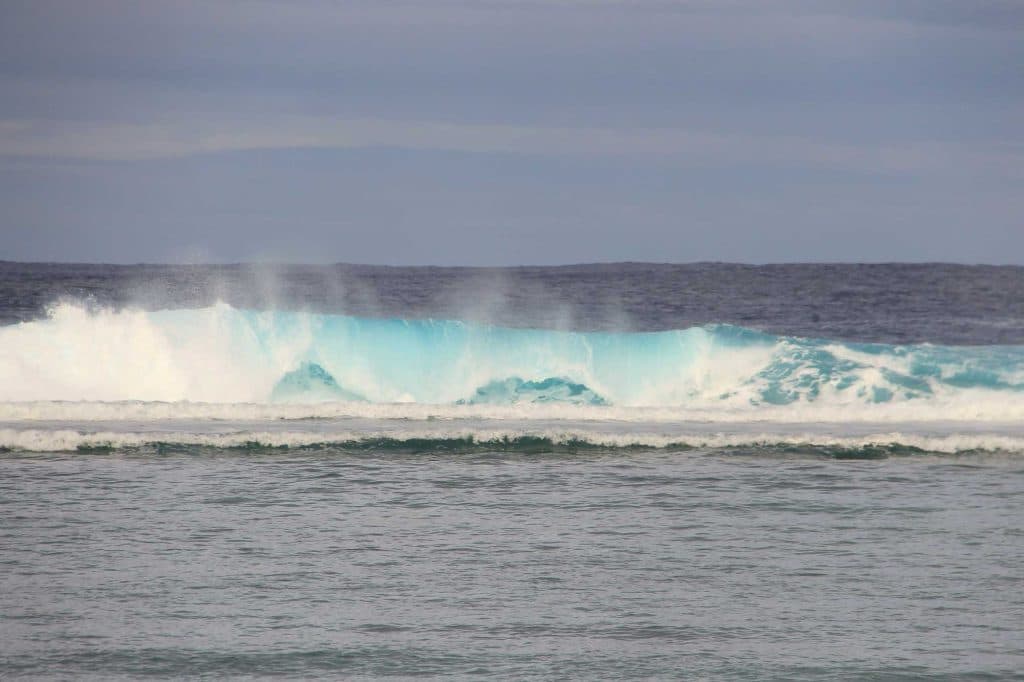 Oceaangolven botsen tegen elkaar onder een bewolkte hemel, met wit schuim en blauw water dat afsteekt tegen de donkere zee, wat de wilde schoonheid van de Cookeilanden oproept.