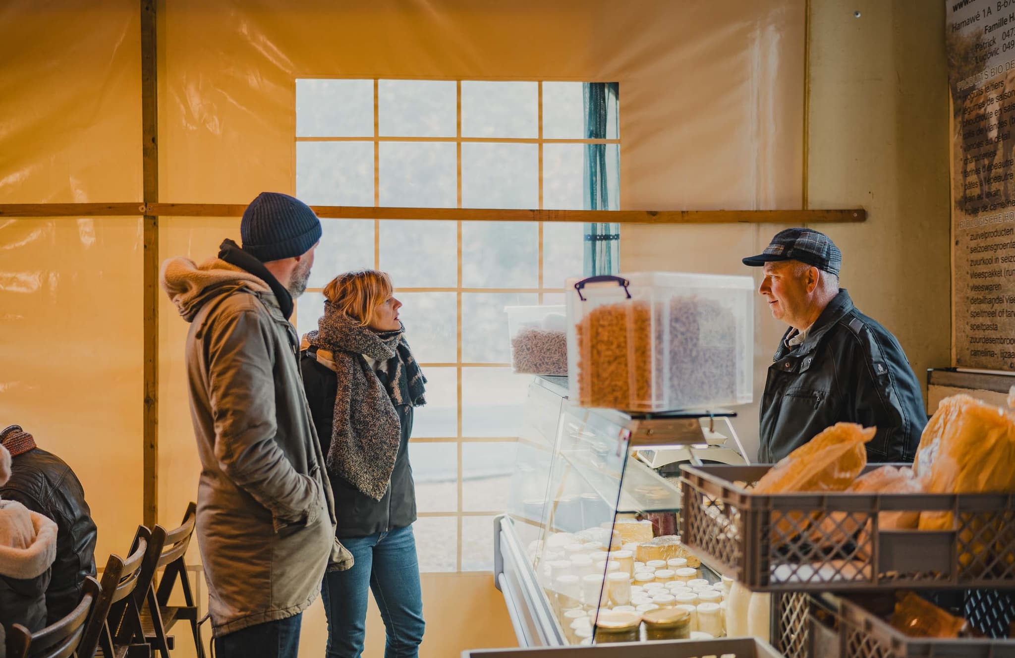 Een man en vrouw in winterkleding staan bij een marktkraam en praten met een verkoper achter een toonbank met kaas en bakjes noten of granen - een charmant tafereel dat een reisblog over lokale hoogtepunten waardig is. Zacht daglicht valt door het grote raam achter hen.