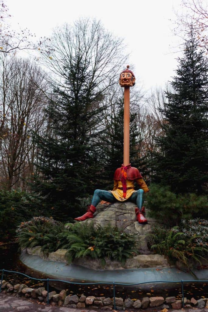Een beeld van een zittende figuur met een lange nek en een hoofd aan de bovenkant, omringd door bomen en groen in een parkachtige omgeving in de Efteling. De figuur draagt kleurrijke kleding en rode laarzen - een must-see voor iedereen die zich afvraagt wat te doen in de Efteling.