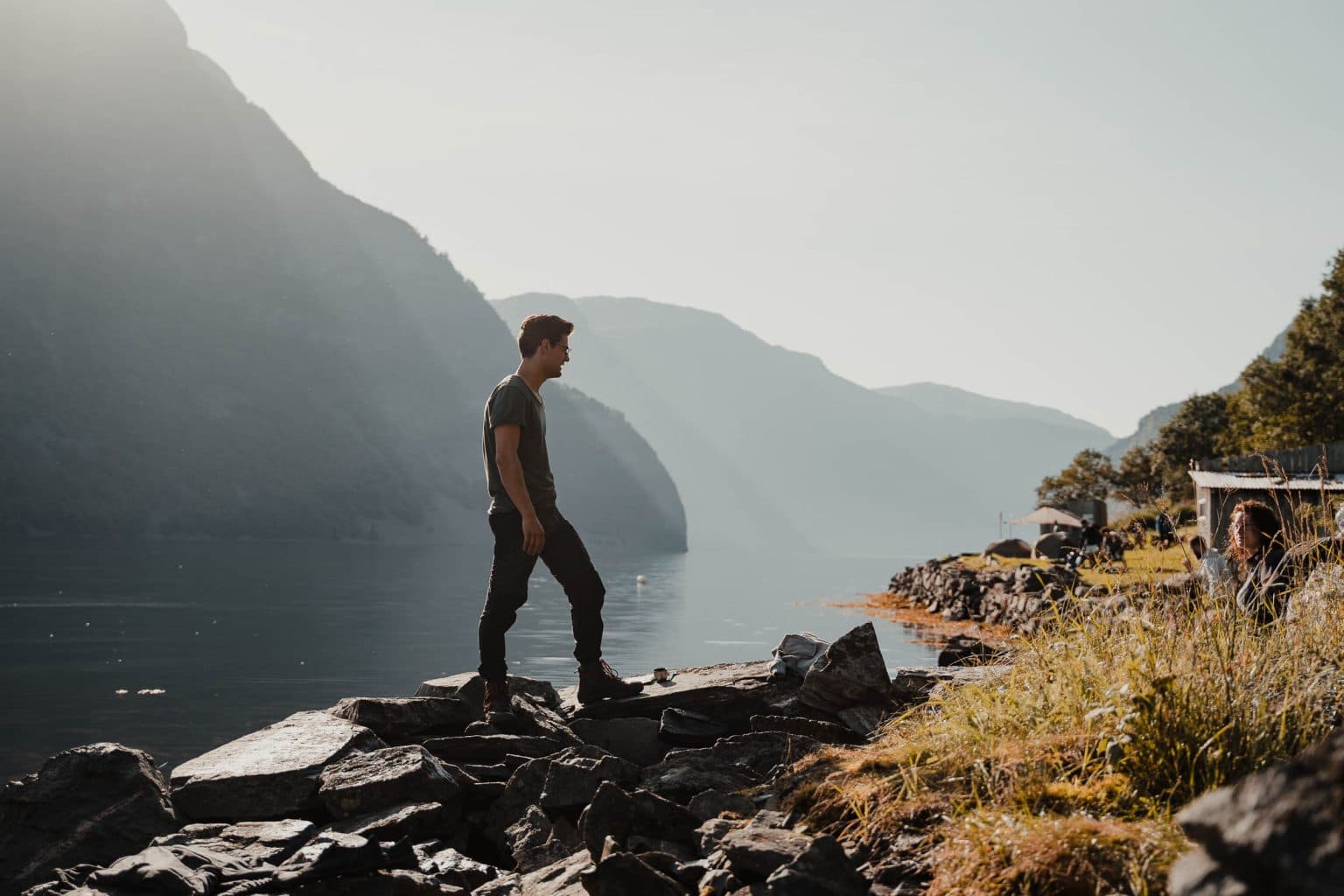 Een persoon staat op een rotsachtig terrein bij een rustig meer, omringd door hoge bergen onder een heldere hemel. Zonlicht creëert schaduwen en highlights, en er groeit groen aan de rand van het water - de perfecte inspiratie voor je volgende reisblog of nieuwe reistips.