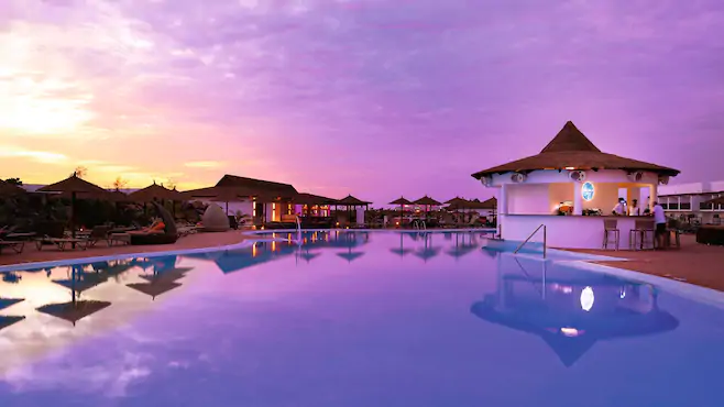 Een rustig buitenzwembad in Kaapverdie met een swim-up bar wordt getoond bij zonsondergang onder een paarse en oranje lucht, omringd door loungestoelen, parasols en zachte verlichting van nabijgelegen gebouwen.
