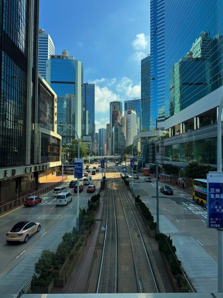 Een straatbeeld met licht verkeer, tramsporen in het midden en hoge glazen wolkenkrabbers aan weerszijden onder een blauwe lucht met verspreide wolken - een van de hoogtepunten en ideaal voor reistips.