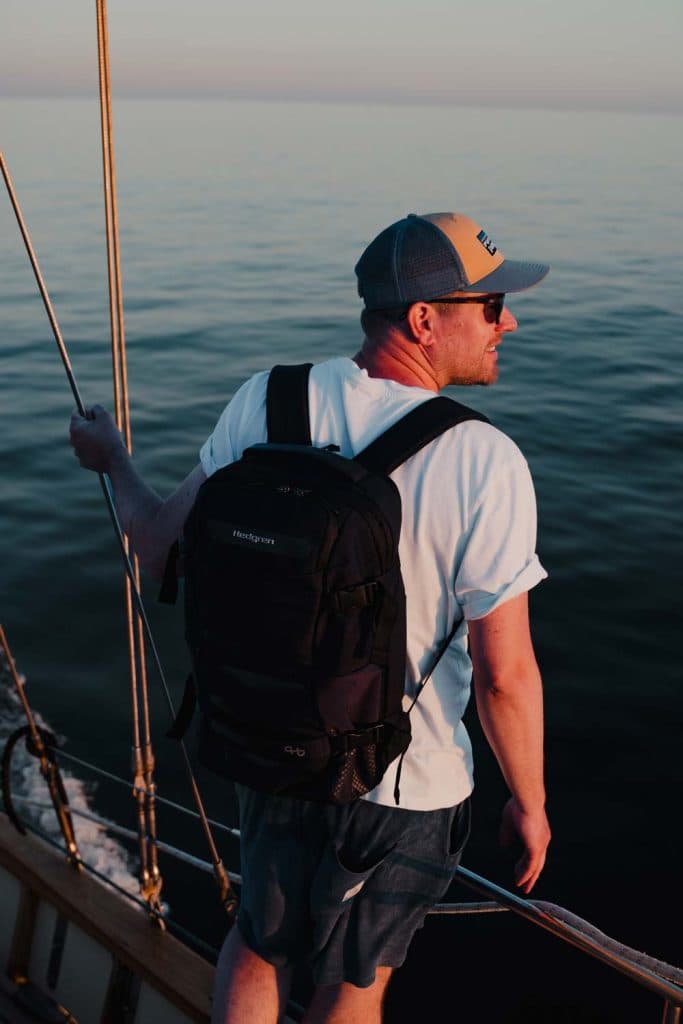 Een man met pet, zonnebril, rugzak en wit T-shirt staat op een boot in Abu Dhabi, houdt een reling vast en kijkt uit over kalm water tijdens zonsondergang - een van de hoogtepunten die Abu Dhabi te bieden heeft.