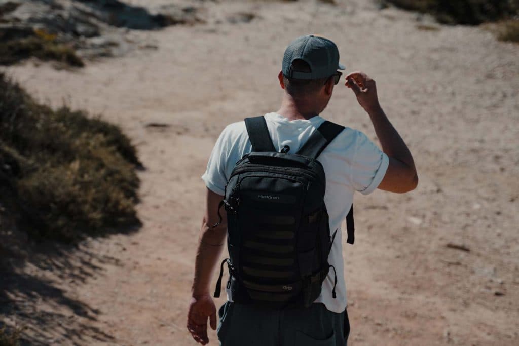 Een persoon met een wit t-shirt, grijze pet en rugzak loopt langs een rotsachtig, zonovergoten pad omringd door schaarse vegetatie in Abu Dhabi en legt de avontuurlijke geest van hoogtepunten Abu Dhabi van achteren vast.