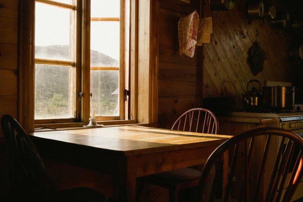 Zonlicht stroomt door een raam naar een houten eettafel met stoelen in een gezellige, rustieke keuken - perfect om verhalen te delen na een dag wandelen of hiken in de schilderachtige landschappen van Noorwegen. Potten hangen aan een wand met houten panelen, met een fornuis op de achtergrond.