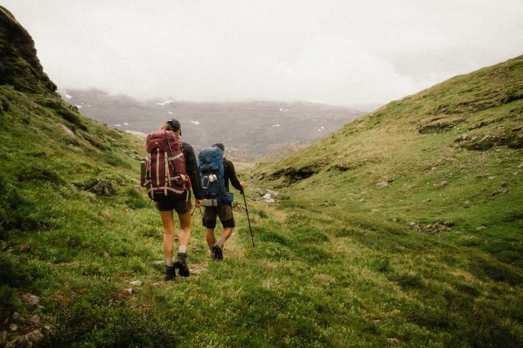 Twee wandelaars met rugzakken lopen over een smal, grassig pad tussen groene heuvels op een bewolkte dag, omringd door bergen en stukken sneeuw in de verte - perfect om te wandelen in Noorwegen.
