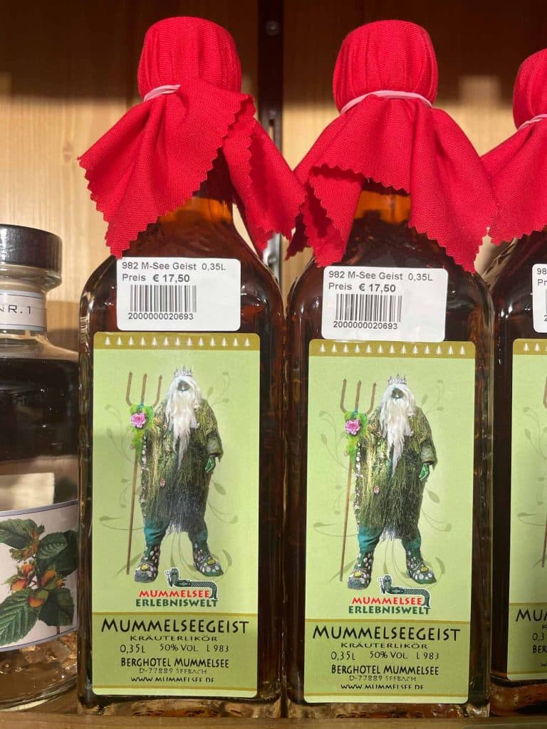 Twee flessen "Mummelseegeist" gedistilleerde drank van het Zwarte Woud, met rode stoffen hoezen en prijsstickers van € 17,50. Op het etiket staat een bebaarde, geklede figuur met een drietand. Een glazen fles is gedeeltelijk zichtbaar aan de linkerkant - perfect na het wandelen in het Zwarte Woud!.