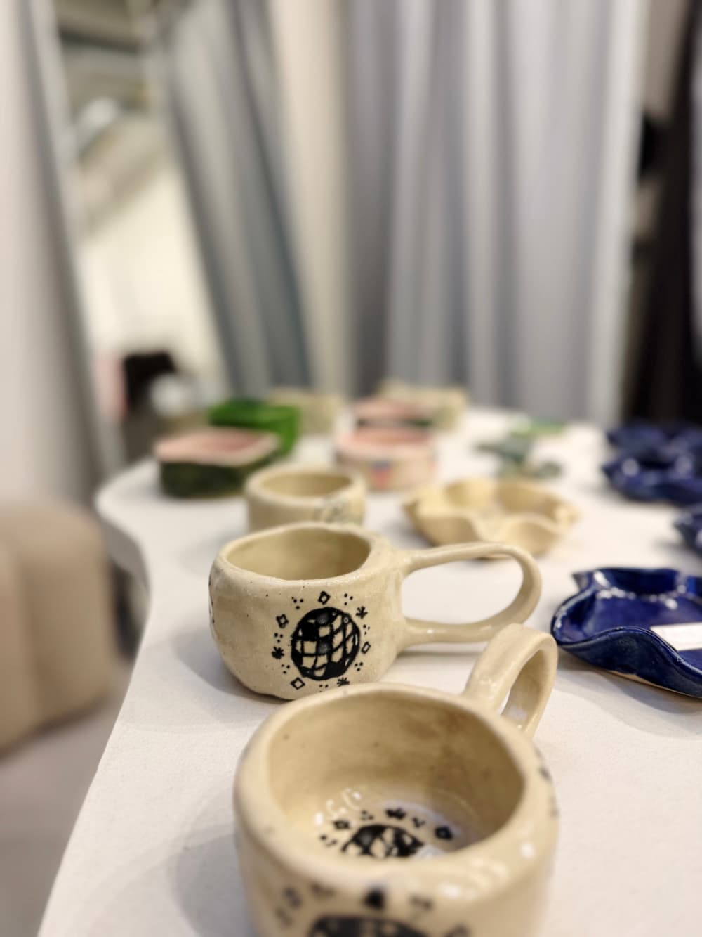 Handgemaakte keramische mokken en kleine schaaltjes, waaronder beige kopjes met zwarte motieven en blauwe en groene stukken, staan uitgestald op een witte tafel - perfect voor liefhebbers van vintage shoppen in Hasselt.