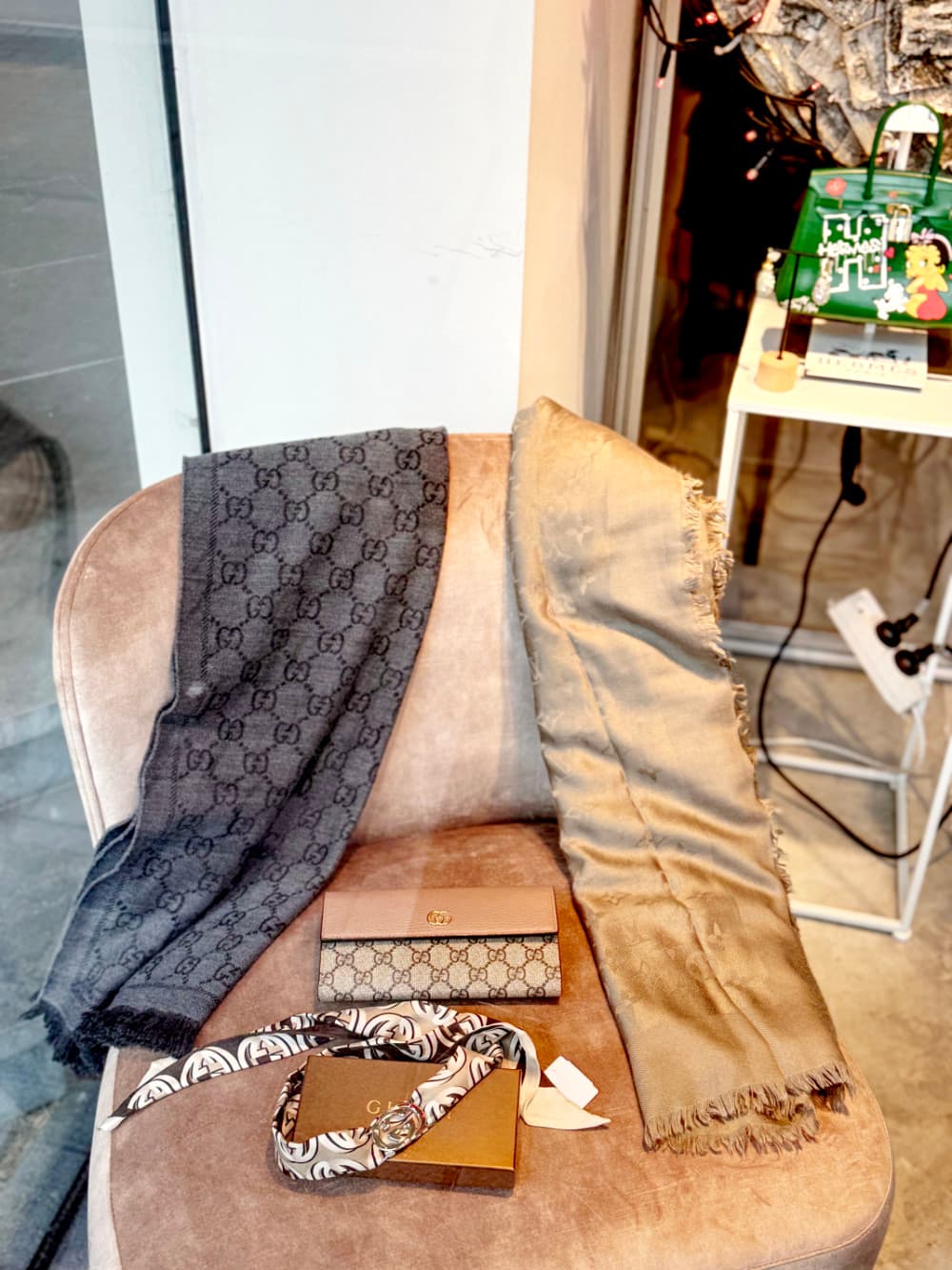 In een beige fluwelen stoel bij het raam liggen een sjaal met zwart patroon, een sjaal met gouden franjes, een bruin Gucci-doosje, een lint met patroon en een kleine Gucci-portemonnee - perfecte vondsten voor vintage shoppen in Hasselt. Een groene tas staat op een plank op de achtergrond.