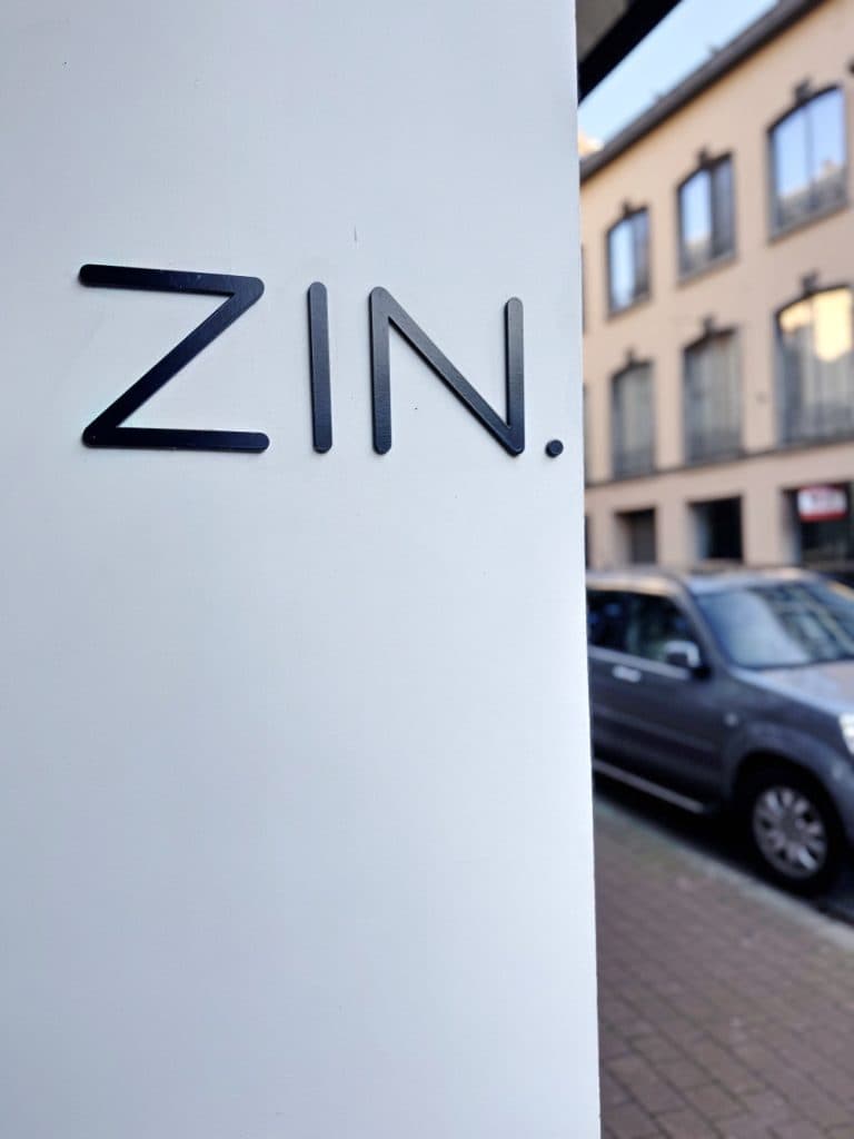 Een witte muur met het woord "ZIN." in zwarte letters; op de achtergrond zijn een geparkeerde auto en een gebouw met grote ramen zichtbaar langs een stadsstraat, die de vibe van vintage shoppen in Hasselt vastleggen.