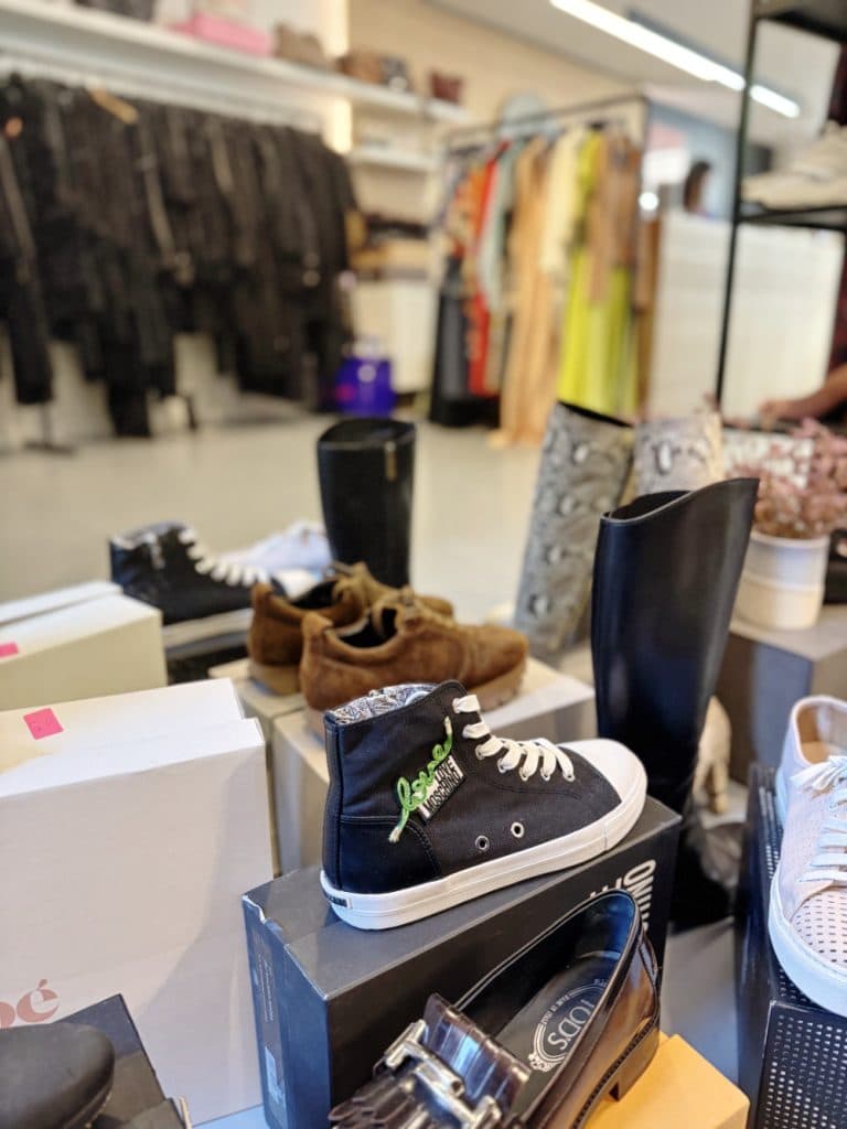 Een uitstalling van verschillende schoenen, waaronder zwarte high-top sneakers en bruine loafers, is gerangschikt op dozen in een kledingwinkel - perfect voor wie wil gaan vintage shoppen in Hasselt, met rekken met kleding zichtbaar op de achtergrond.