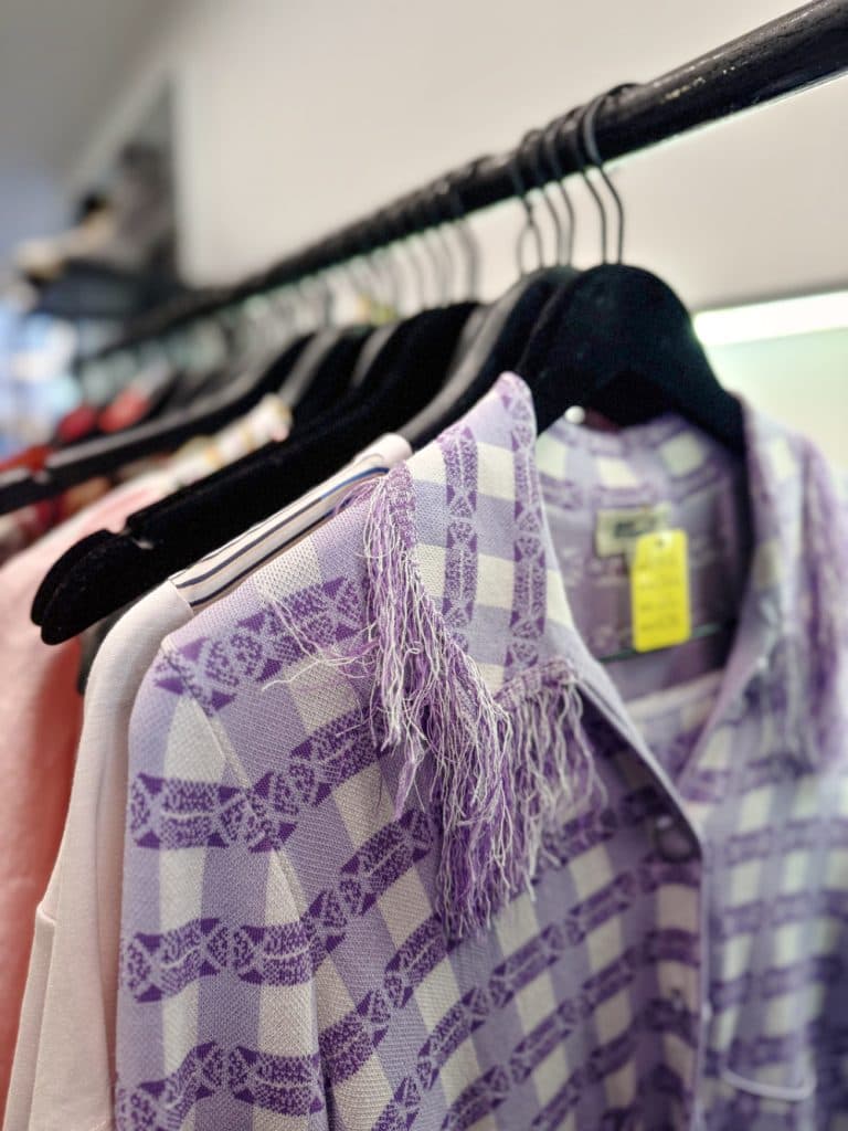 Close-up van een lavendelwit geruit hemd met gerafelde draden op de schouder, hangend op een zwarte hanger in een kledingwinkel in Hasselt, perfect voor iedereen die houdt van vintage shoppen in Hasselt, omringd door andere unieke kledingstukken.