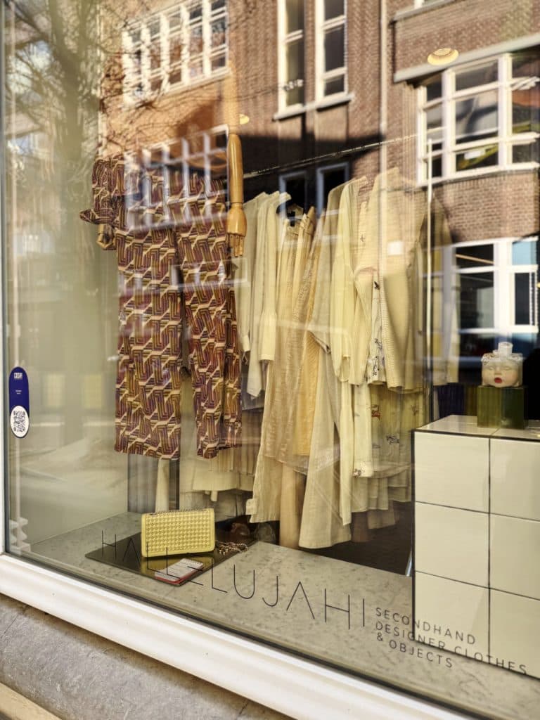 Etalage in Hasselt met gedessineerde en effen crèmekleurige tweedehands jurken op hangers, een gele handtas op een witte kubus en weerspiegelingen van bakstenen gebouwen - perfect voor vintage shoppen in Hasselt.
