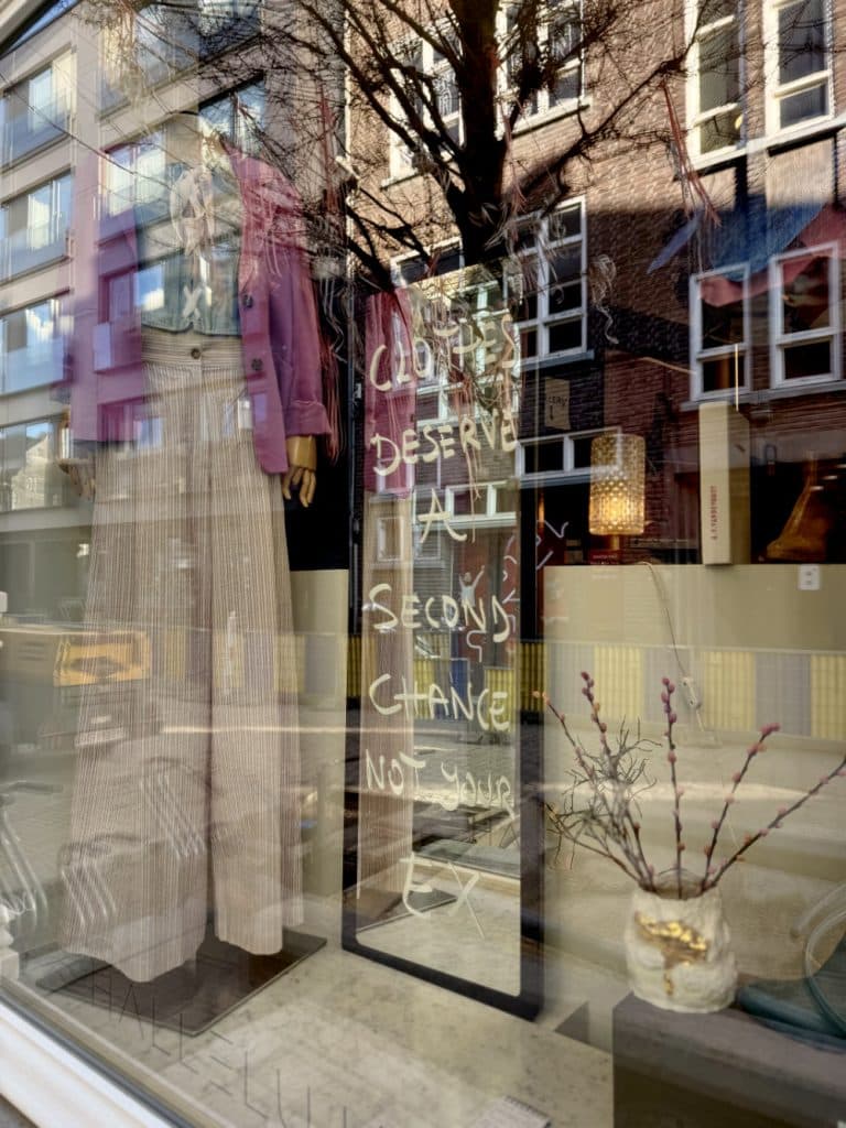 Een etalage in Hasselt toont mannequins gekleed in stijlvolle kleding en een spiegel met handgeschreven tekst: "Kleren verdienen een tweede kans, niet je ex." Reflecties van gebouwen en bomen onthullen de vintage shoppen in Hasselt vibe.