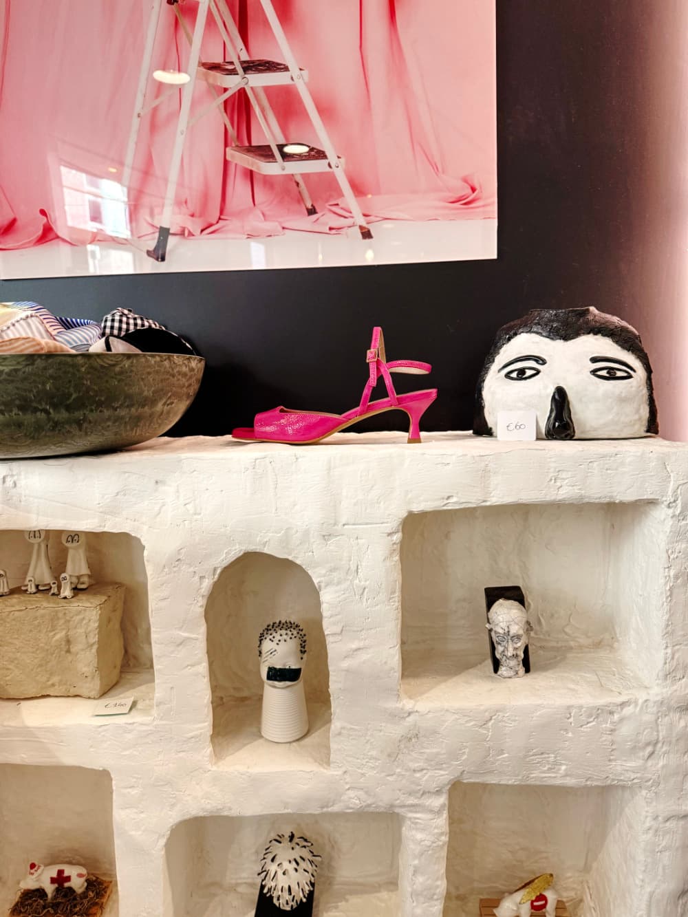 Op een witte displayplank staan decoratieve voorwerpen, waaronder een zwart-wit gezichtssculptuur en een roze sandaal met hoge hakken - de perfecte inspiratie voor liefhebbers van vintage shoppen in Hasselt. Boven hangt een foto van een roze ladder aan een donkere muur.