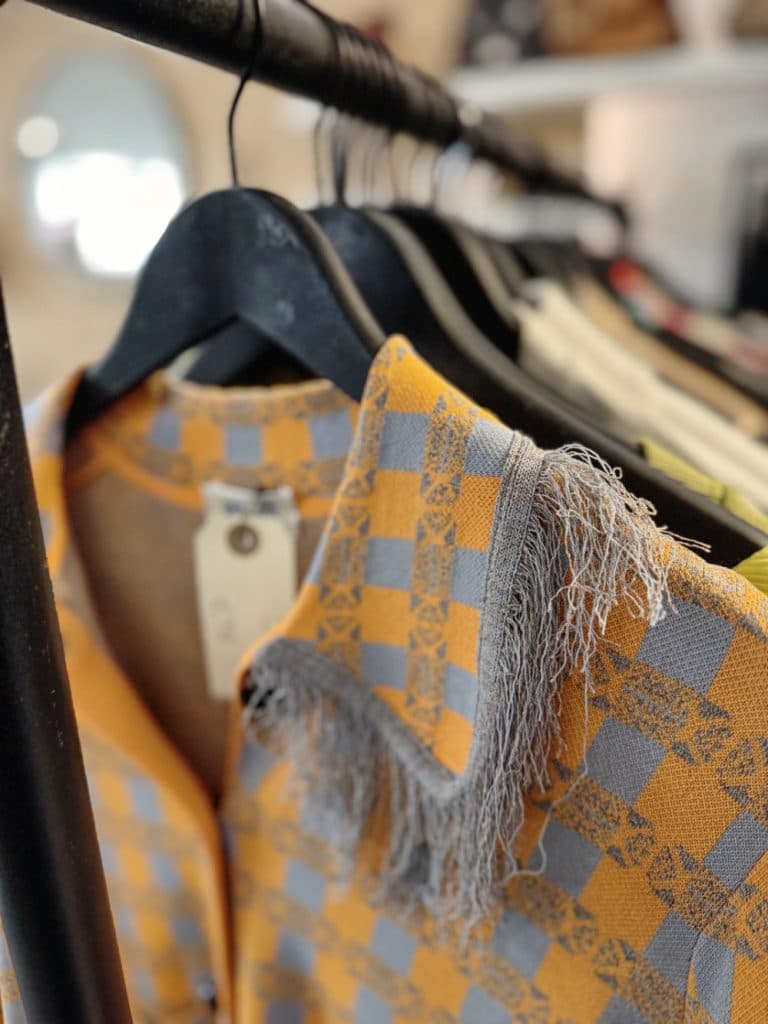 Een close-up van een geel en grijs geruit overhemd met een gerafelde kraag hangend aan een zwarte hanger, uitgestald tussen andere kledingstukken op een rek - perfect voor vintage shoppen in Hasselt.