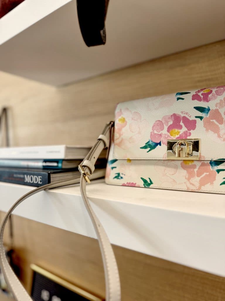 Een handtas met bloemmotief en een beige riem rust op een witte plank naast boeken - een ideale vondst voor liefhebbers van vintage shoppen in Hasselt. De scène is voorzien van lichte houten lambrisering en een glimp van andere planken.