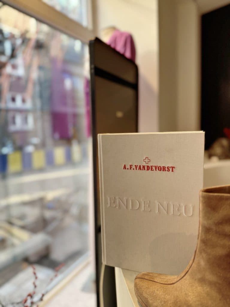 Een boek met de titel "ENDE NEU" van A.F. VANDEVORST staat rechtop op een oppervlak naast een tan suède laars, wat de charme oproept van een vintage winkel in Hasselt, met een vaag straatbeeld door een groot raam op de achtergrond.