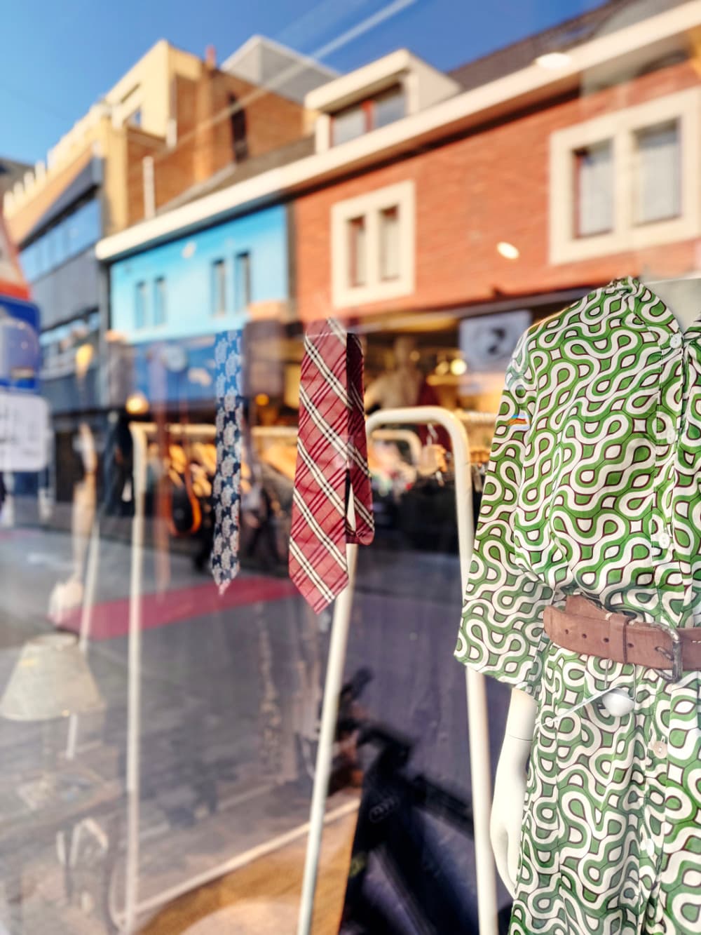 Een etalage in Hasselt met een groen-wit gedessineerde jurk met een bruine riem en twee stropdassen, met gebouwen en straatreflecties op het glas - perfect voor wie houdt van vintage shoppen in Hasselt.