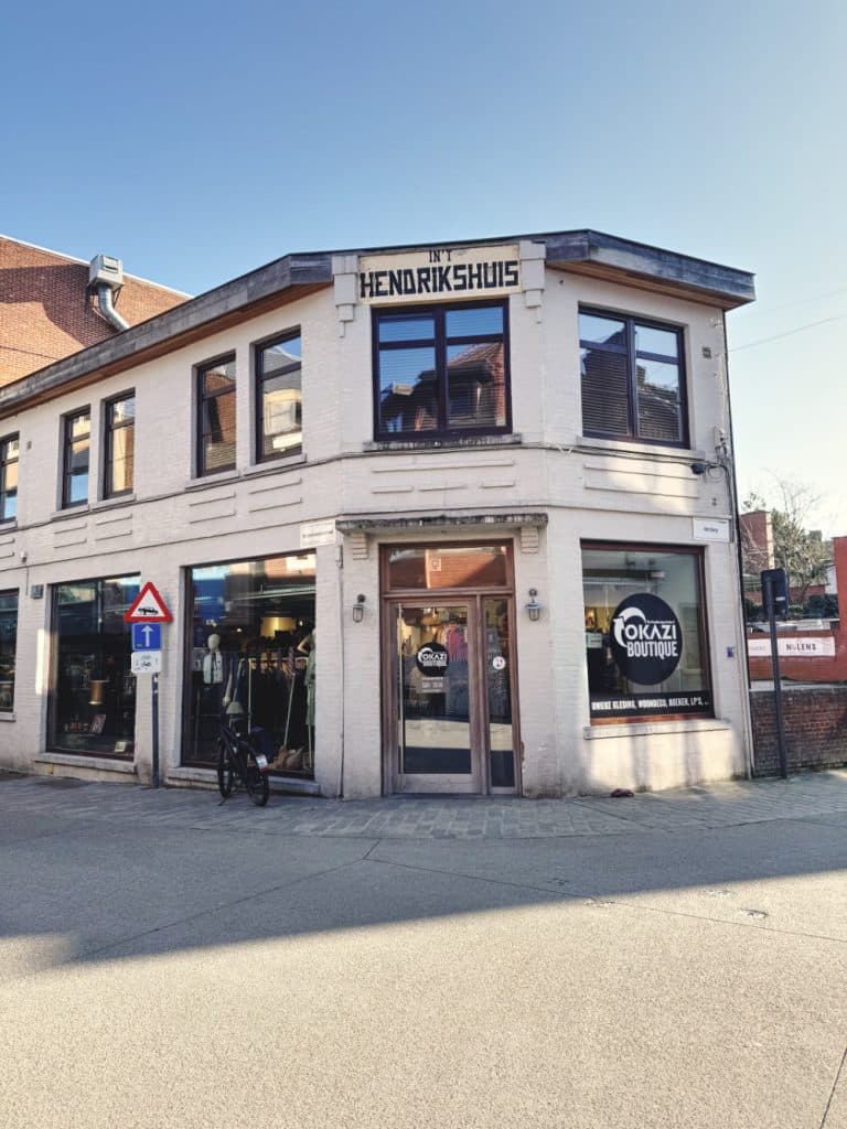 Een bakstenen hoekpand met grote ramen en "Hendrikshuis" erop geschreven in Hasselt. Op de begane grond vind je de boetiek "OKAZI", perfect voor vintage shoppen in Hasselt. Buiten staat een fiets geparkeerd en links is een straatnaambordje te zien.