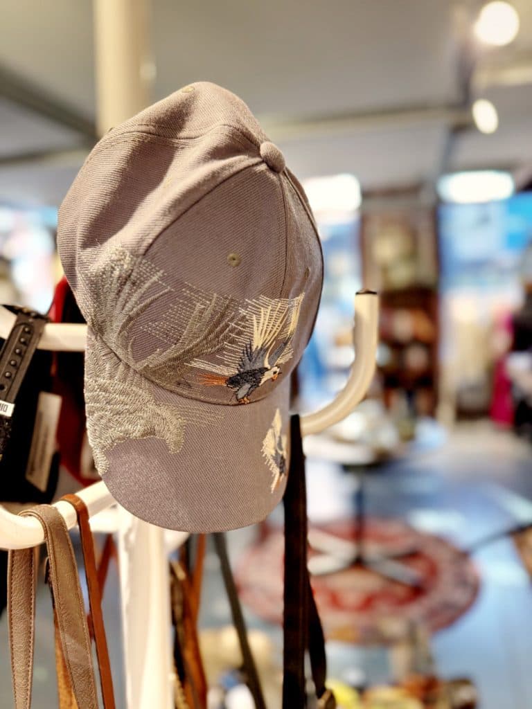 Een grijze baseballpet met geborduurde vogels en gebladerte hangt aan een witte kapstok in een zacht verlichte, gezellige kamer-perfecte inspiratie voor vintage shoppen in Hasselt, met een rond gedessineerd tapijt en vaag meubilair op de achtergrond.